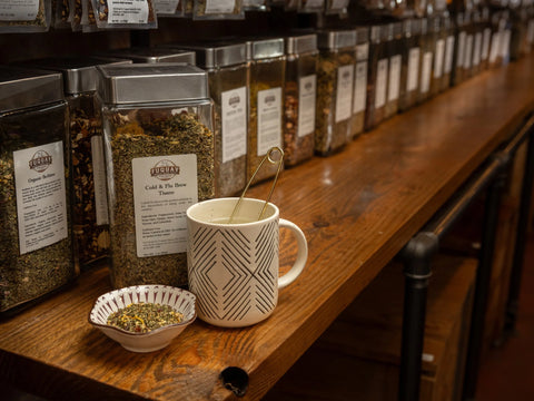 Glass jars of herbs and tea displayed on a wooden shelf with a cup of tea.